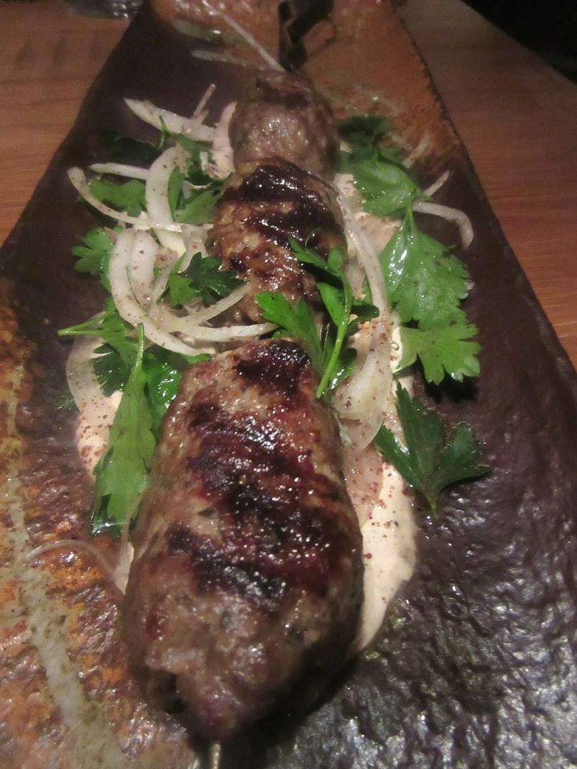 Wood grilled lamb kebab is served on the skewer on a massive ceramic plate. The server removes the skewer.